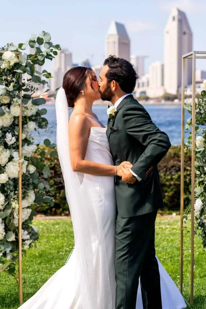 Newlyweds kiss under floral arch with San Diego skyline in the background Coronado Centennial Park