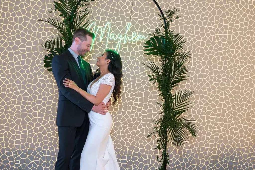 Bride and groom pose under a neon sign and greenery arch at modern San Diego Skybox wedding