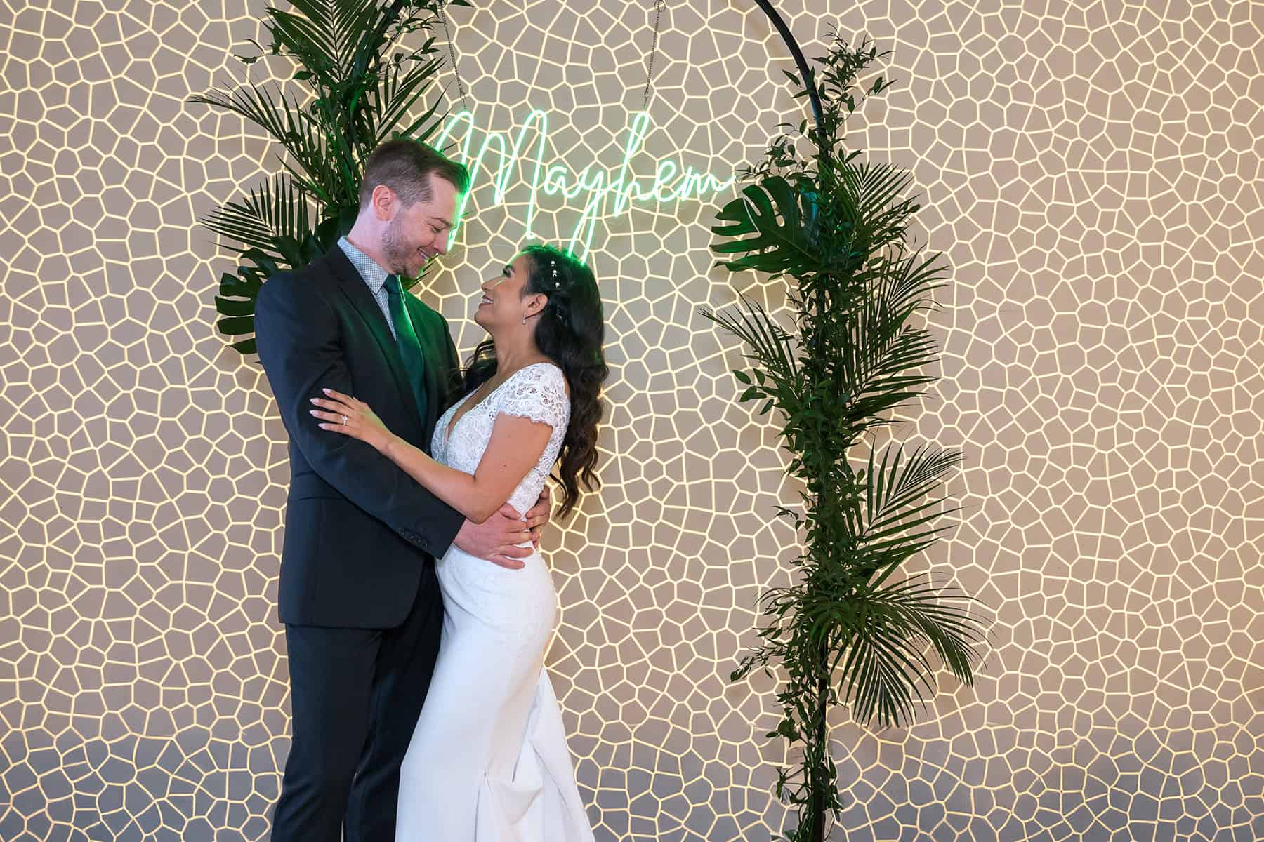 modern-wedding-bride-groom-neon-arch-san-diego Bride and groom pose under a neon sign and greenery arch at modern San Diego Skybox wedding