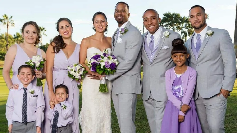 Wedding party of nine posing outdoors in San Diego with bride and groom, bridesmaids in lavender dresses, groomsmen in gray suits, and children, holding white and purple floral bouquets against green grass and palm trees.