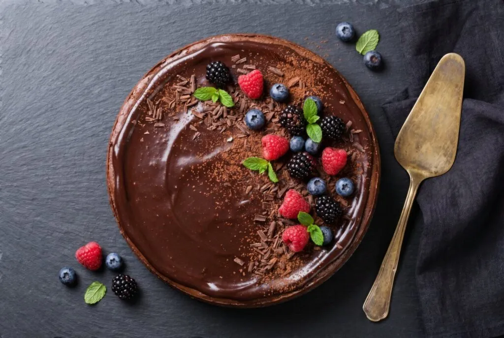 Rich chocolate layer cake decorated with fresh raspberries, blackberries, blueberries, and mint leaves on dark slate surface with gold cake server