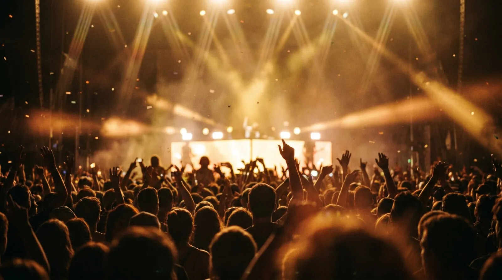 Large crowd with raised hands at concert or event with dramatic golden stage lighting and haze, capturing live event energy.