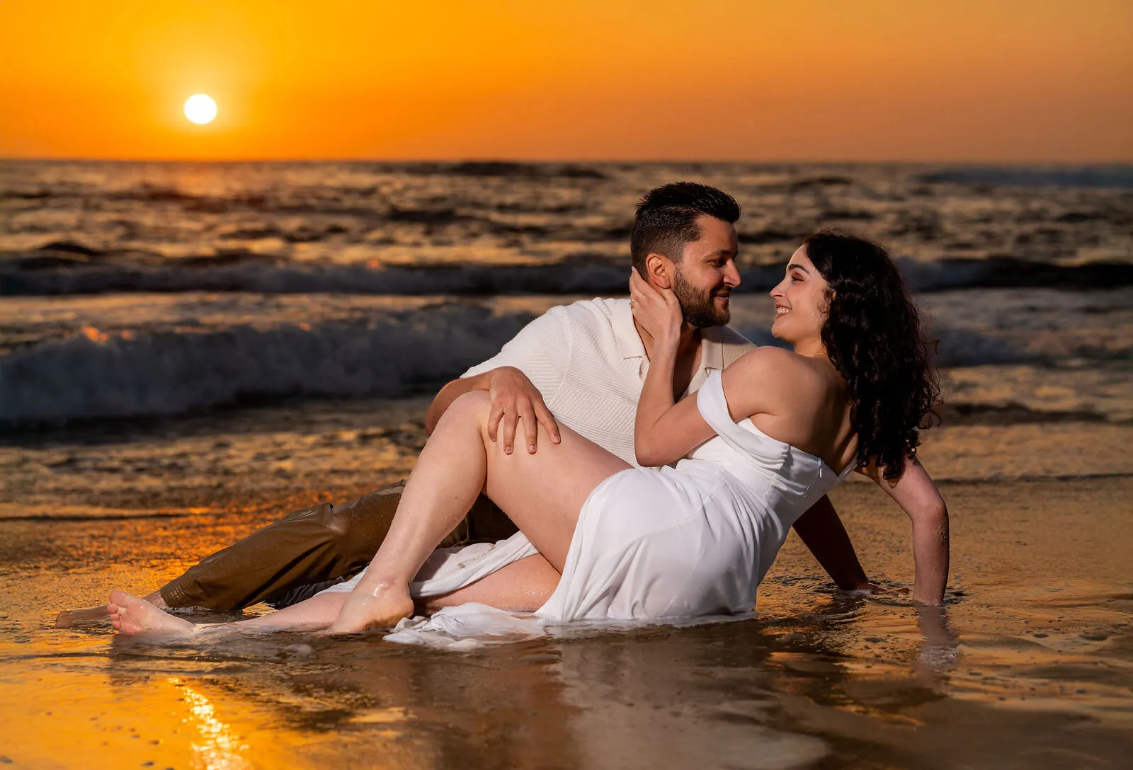 Couple in white clothing posing together on sandy beach during golden hour sunset for engagement photography session.