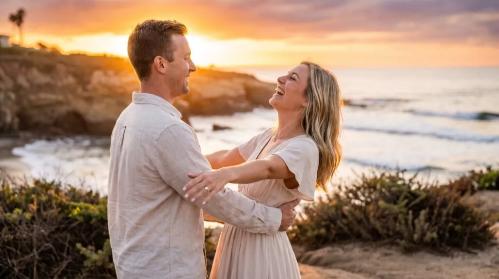 Happy couple embracing and laughing together during a surprise proposal at sunset on a rocky San Diego beach coast.