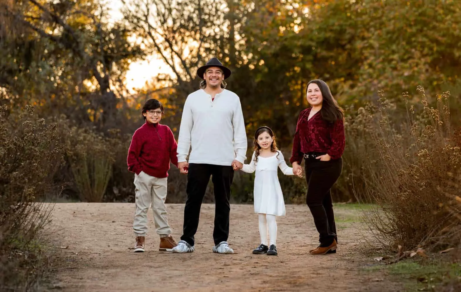 Happy family of four holding hands on a tree-lined autumn path, wearing coordinated burgundy and neutral tones during golden hour.