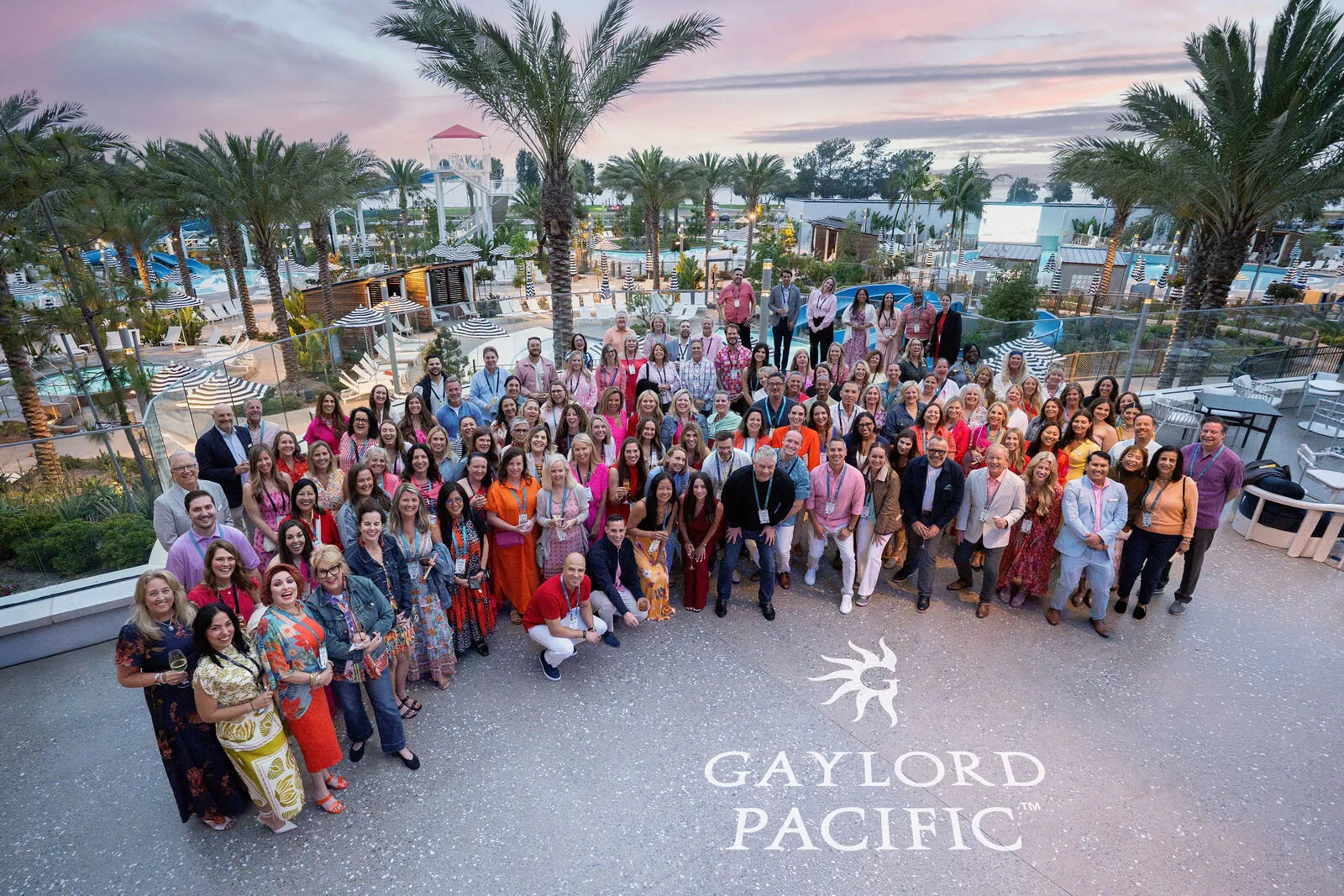 Large group photo of women at Gaylord Pacific resort with palm trees and waterfront backdrop at sunset, capturing a corporate event or conference gathering.