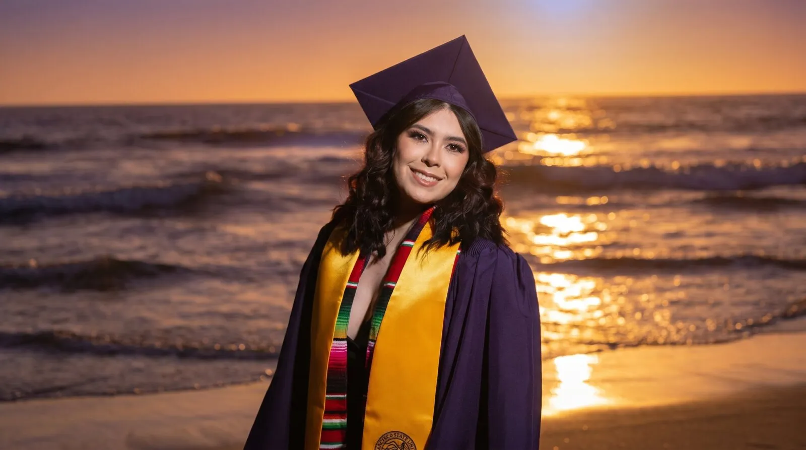 Young woman in purple graduation gown and yellow stole smiles at sunset beach during her graduation milestone celebration in San Diego.