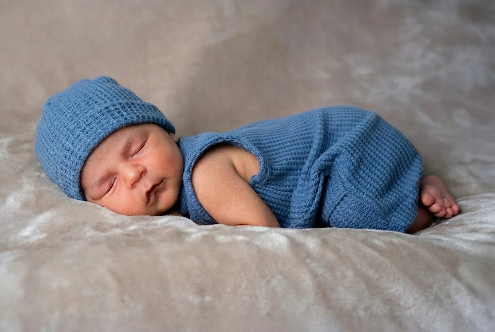 Sleeping newborn baby wearing blue knit outfit and matching hat on soft beige blanket during newborn photography session