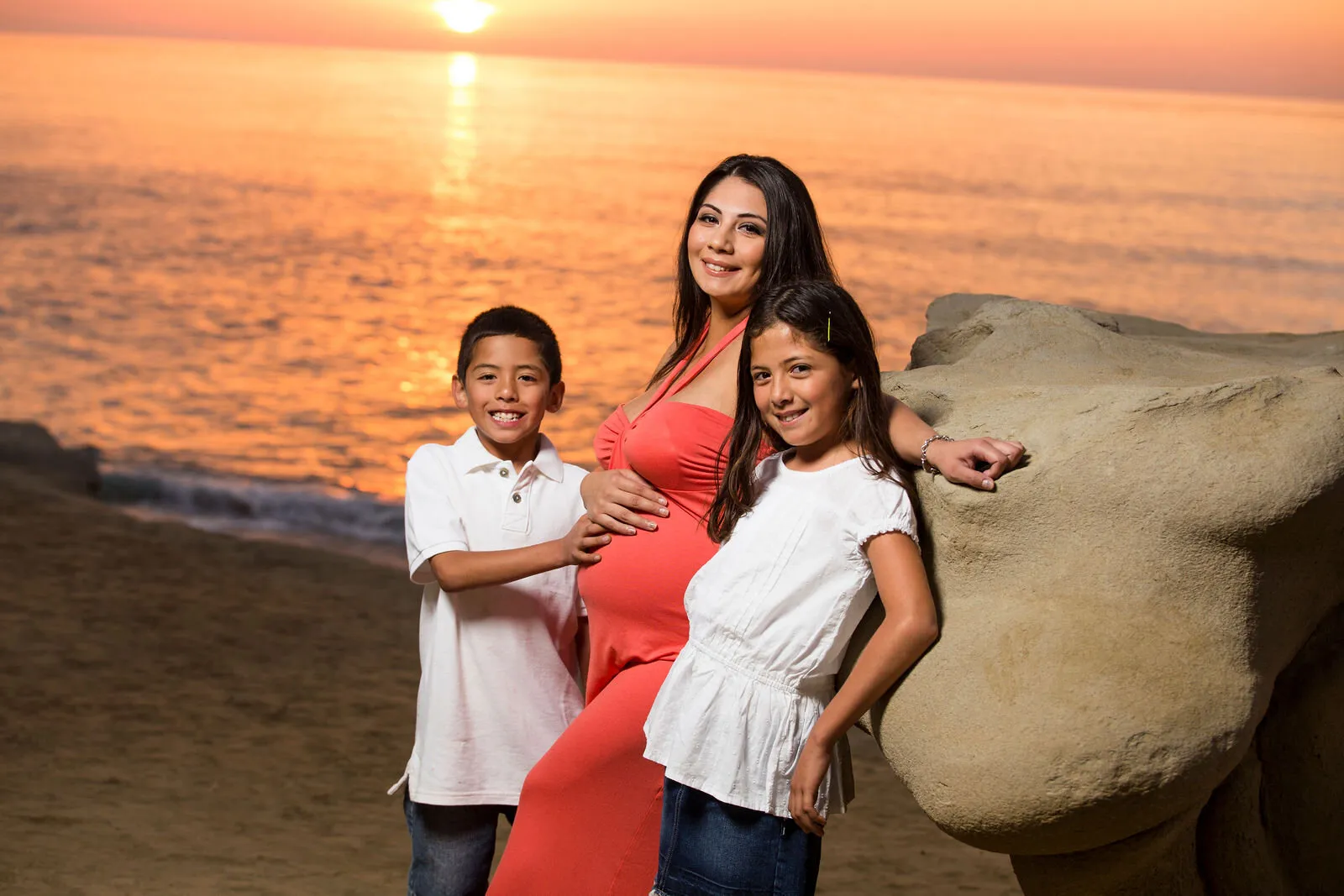 Pregnant mother in coral dress with two children on sandy beach at golden hour sunset, San Diego family maternity portrait