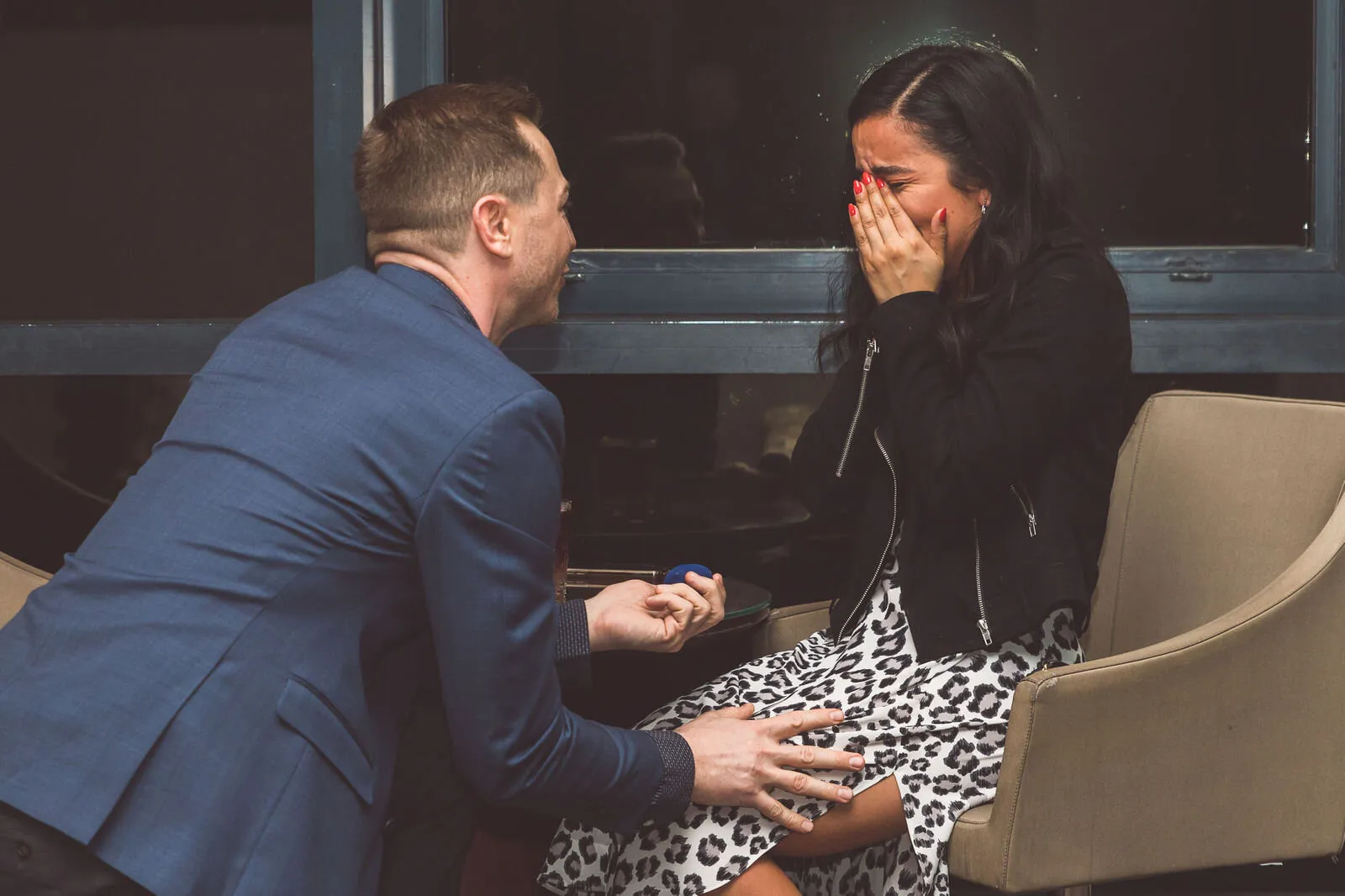 Man in blue suit proposing to woman with engagement ring during emotional proposal moment