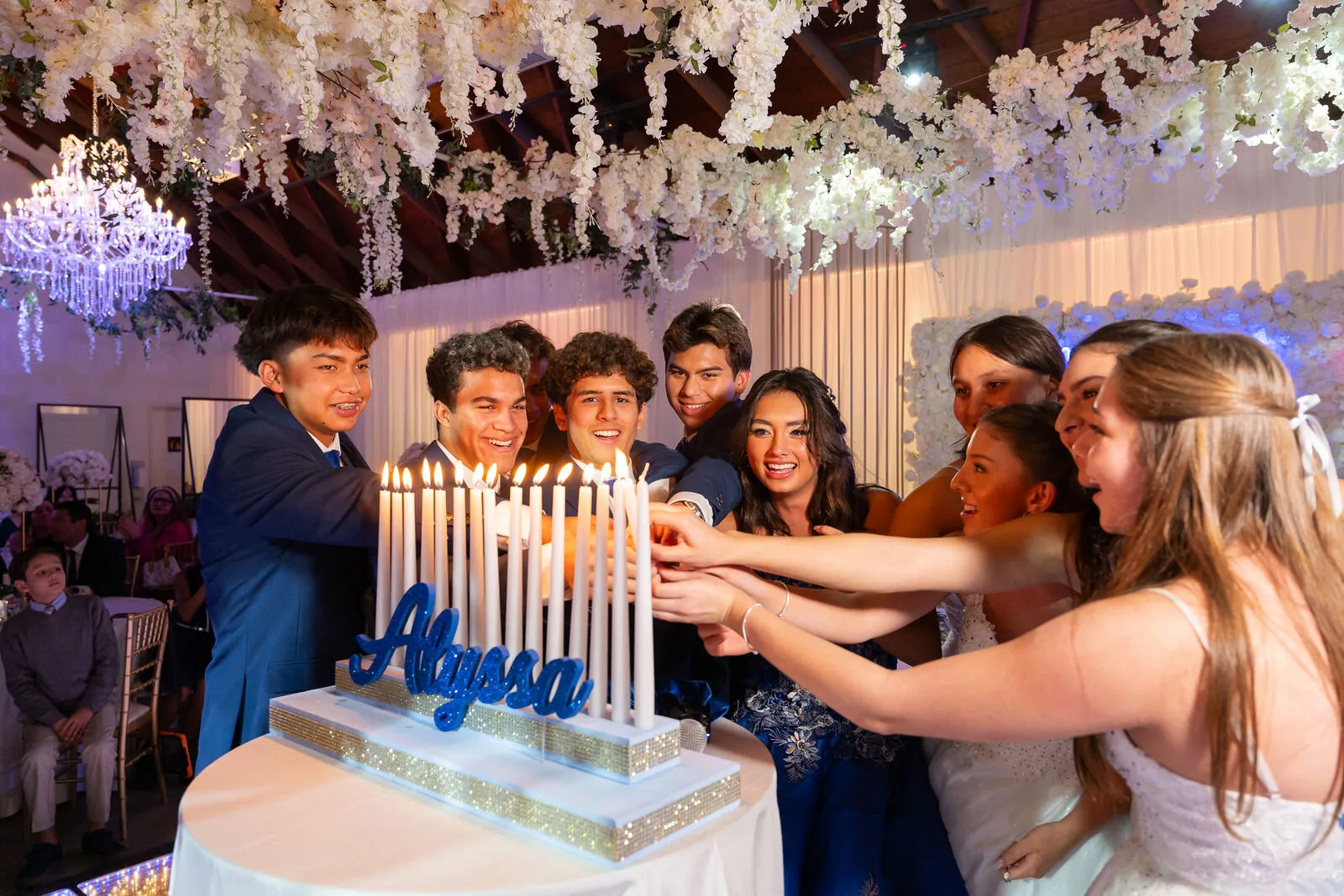 Group of teenagers lighting candles together during a sweet 16 celebration with elegant white floral ceiling decorations and blue lighting.