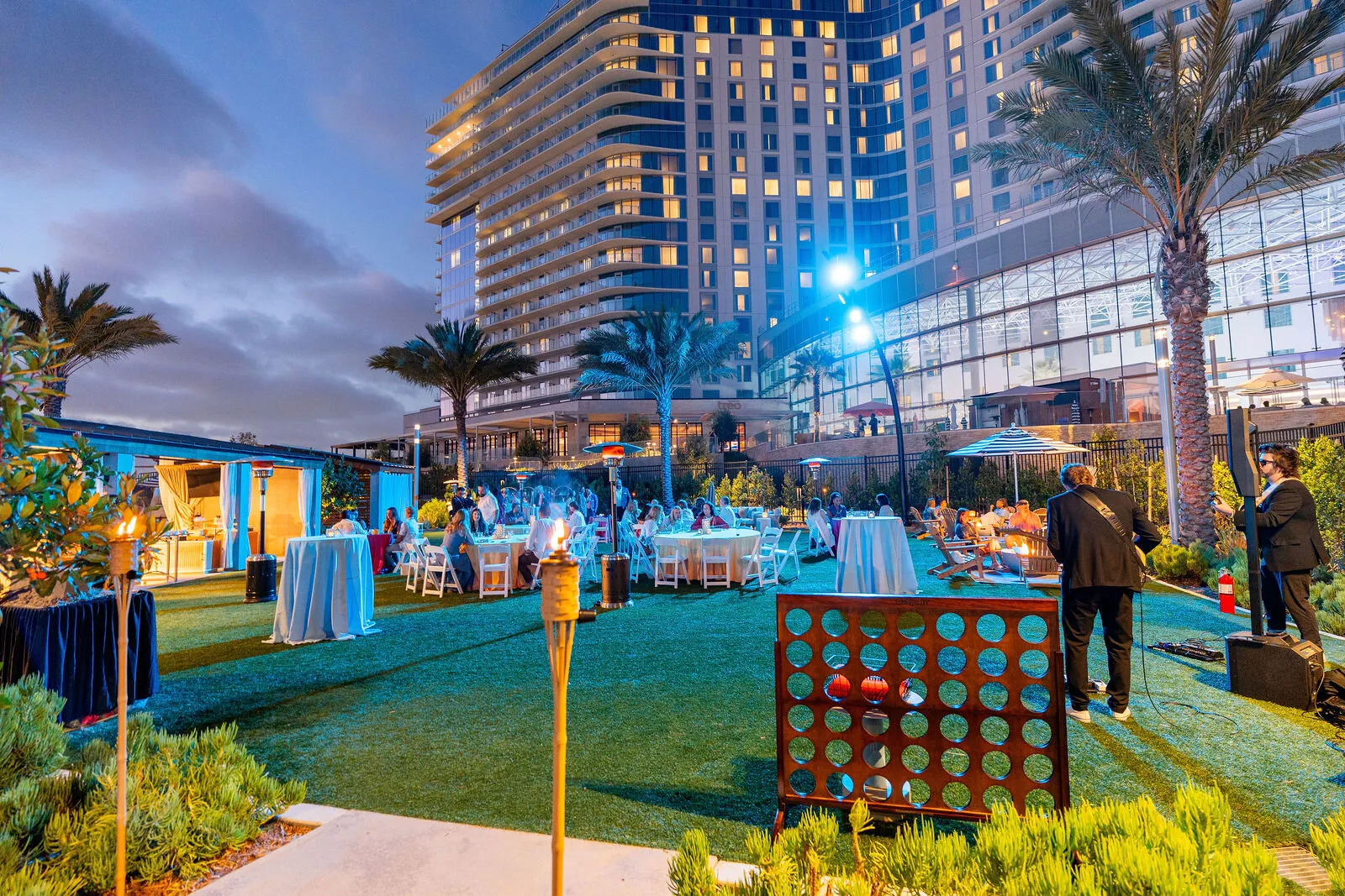 Elegant rooftop wedding reception with blue-lit modern high-rise building, palm trees, and guests at round tables during twilight in San Diego