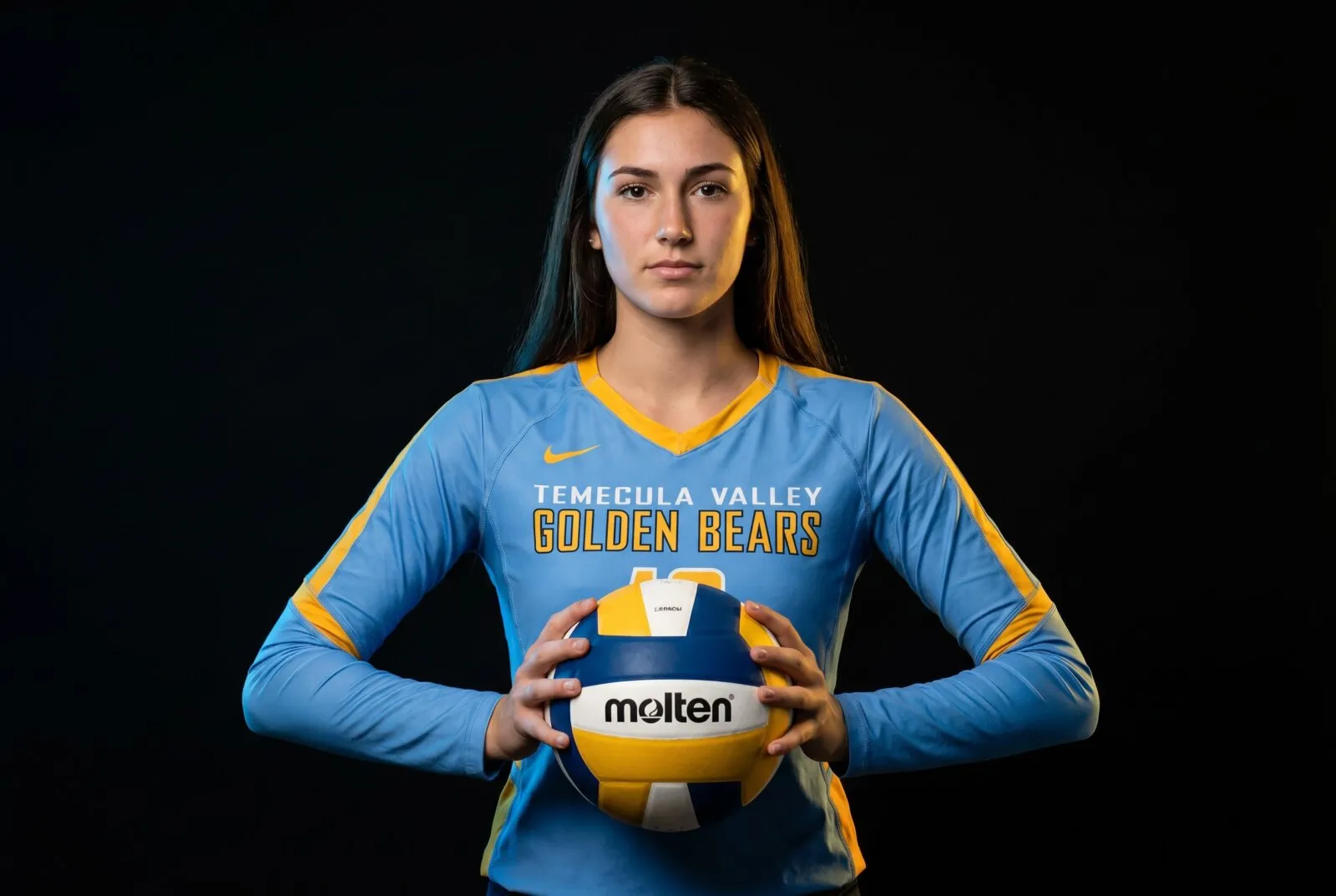 Female volleyball athlete in Temecula Valley Golden Bears blue and gold uniform holding Molten volleyball against black background.