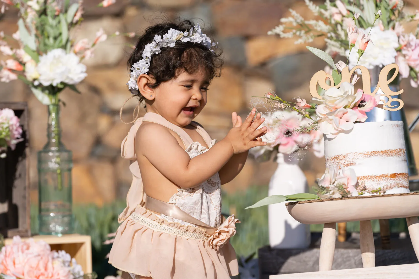 Happy toddler in flower crown reaching toward decorated birthday cake surrounded by pink and white floral arrangements at outdoor celebration.