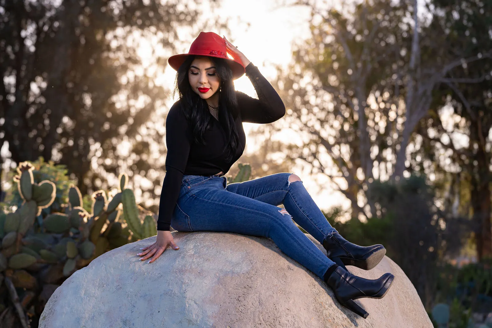 Woman in red hat and blue jeans sits on boulder among cacti in desert landscape, captured by San Diego portrait photographer