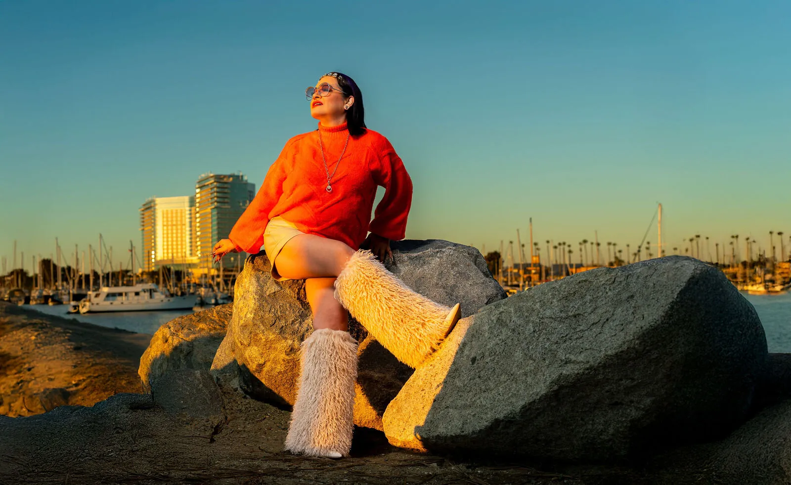 Woman in red sweater sitting on rocks at San Diego beach with boats and marina in background at golden hour